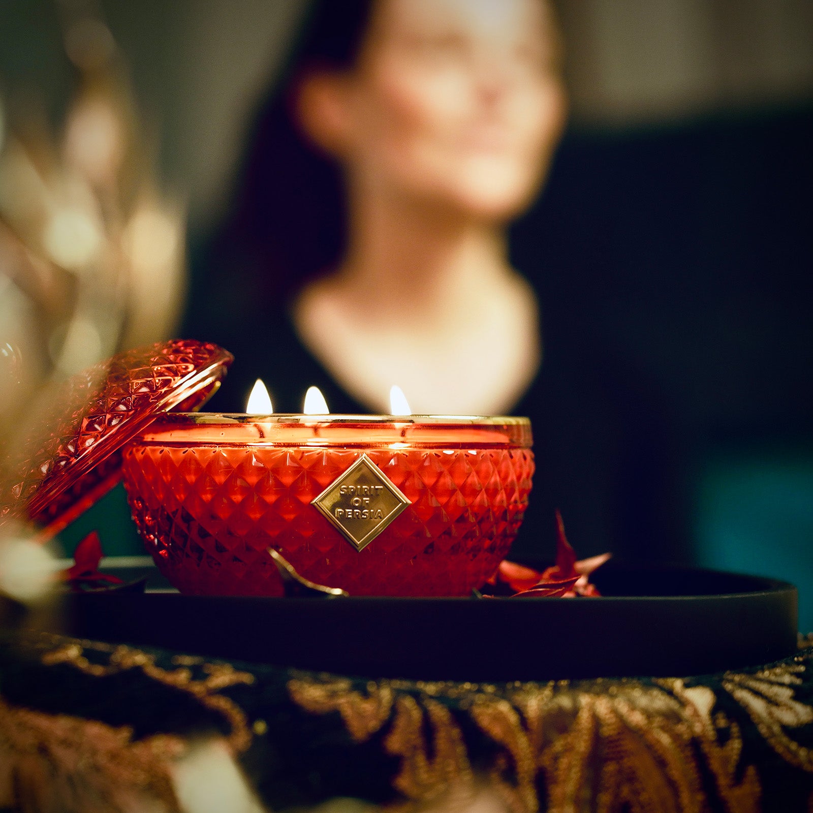 Unsere hochwertige Duftkerze Atussa ist auf einem schwarzen Tablett gestellt. Im Hintergrund meditiert eine dunkelhaarige Frau in einem schwarzen Pullover.