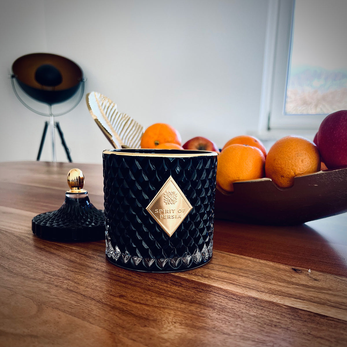 Unsere hochwertige Duftkerze Teheran auf einem Holztisch. Im Hintergrund befindet sich eine goldene Schale mit Orangen und eine schwarz-goldene Stehlampe.