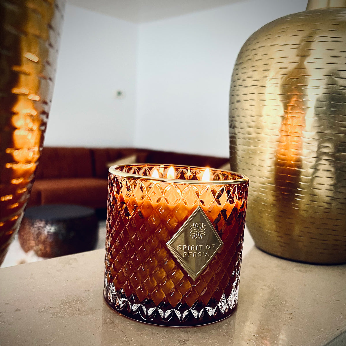 Unsere hochwertige Duftkerze Jazd neben zwei goldenen Vasen. Im Hintergrund befindet sich ein braunes Sofa und ein brauner runder Tisch.