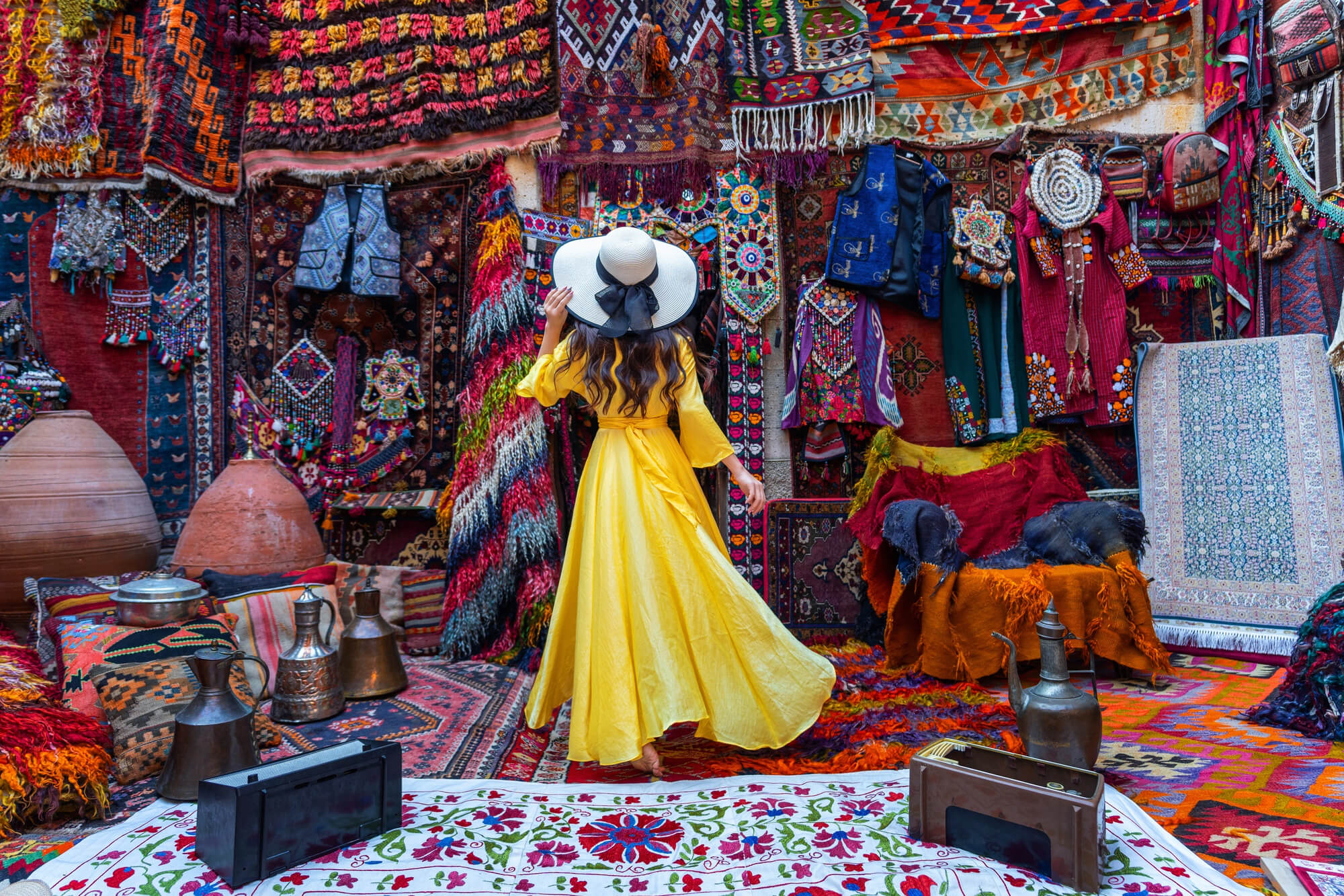 Eine wunderschöne Frau in einem gelben Kleid mit weißem Hut steht in einem persischen Teppichgeschäft und bewundert die handgefertigten orientalischen Produkte.